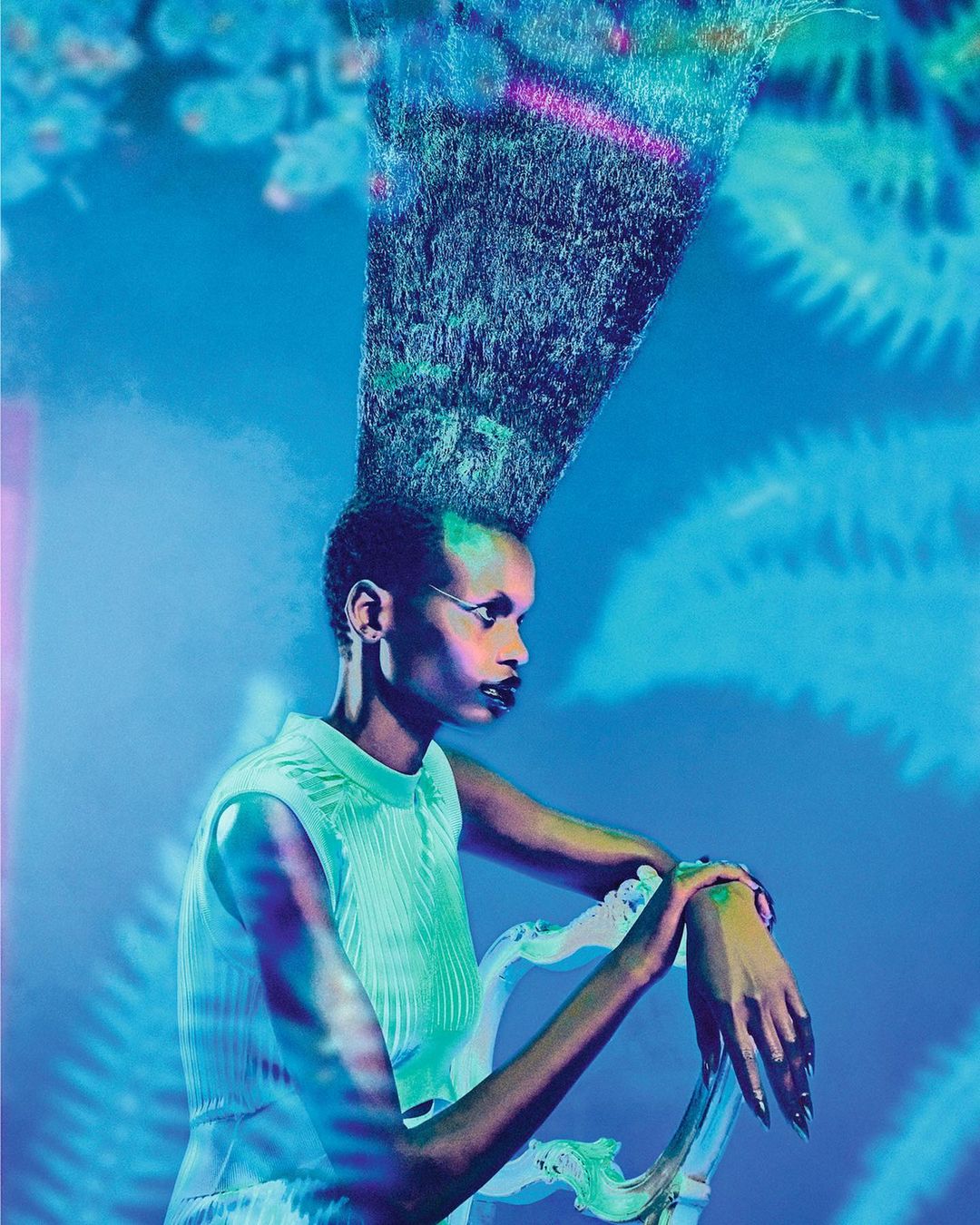 An elegant dark-skinned woman with hair extending upward sits regally on a decorative chair in front of a light blue patterned backdrop.