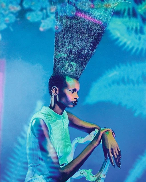 An elegant dark-skinned woman with hair extending upward sits regally on a decorative chair in front of a light blue patterned backdrop.