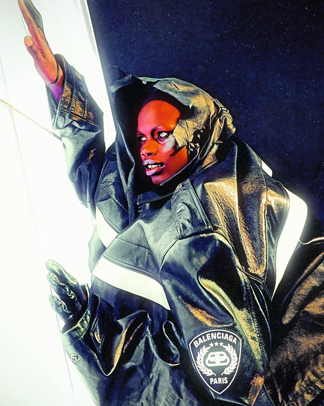 A person in a black hooded leather jacket stands against a dark background, accented by white light. The jacket has a Balenciaga logo.