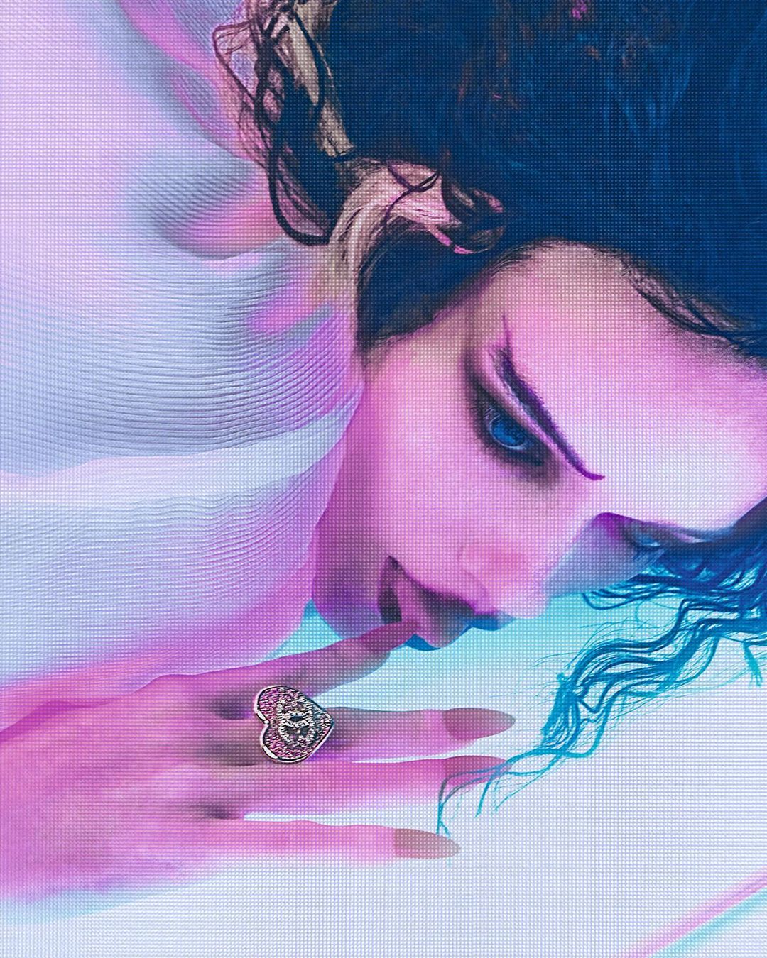 Close-up of a fair-skinned woman with dark hair and blue eyes wearing a heart-shaped ring, lying down, and looking to the side with a soft focus.