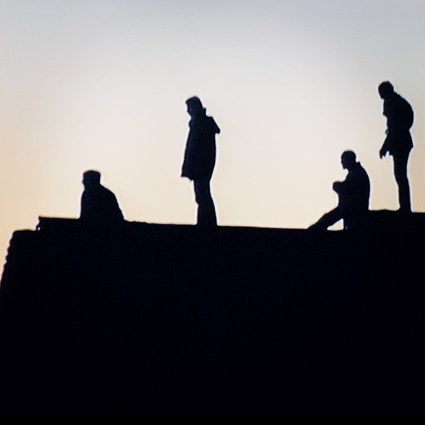 Silhouetted figures stand atop a dark, angular structure against a light sky, creating a stark contrast and minimalist aesthetic.