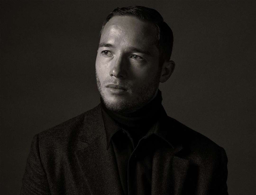 Monochrome portrait of a handsome adult man with short dark hair, wearing a dark turtleneck and blazer, looking off-camera against a dark background.