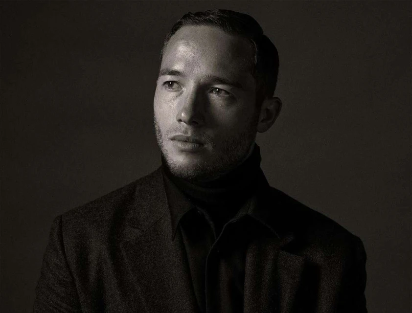 Monochrome portrait of a handsome adult man with short dark hair, wearing a dark turtleneck and blazer, looking off-camera against a dark background.