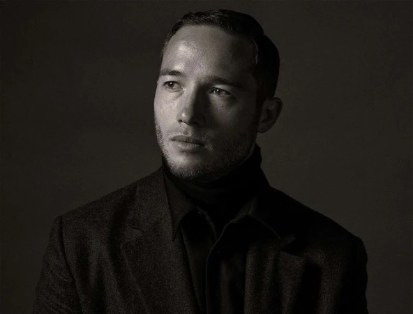 Monochrome portrait of a handsome adult man with short dark hair, wearing a dark turtleneck and blazer, looking off-camera against a dark background.