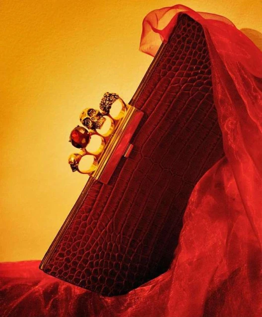 Red clutch purse with crocodile texture and jewel-encrusted knuckle duster handle, presented against a softly draped red fabric and yellow backdrop.