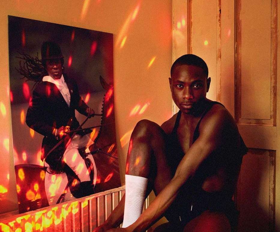 Portrait of a Black man in a tank top sitting in front of a framed image of a person riding a horse, illuminated by red and orange light.