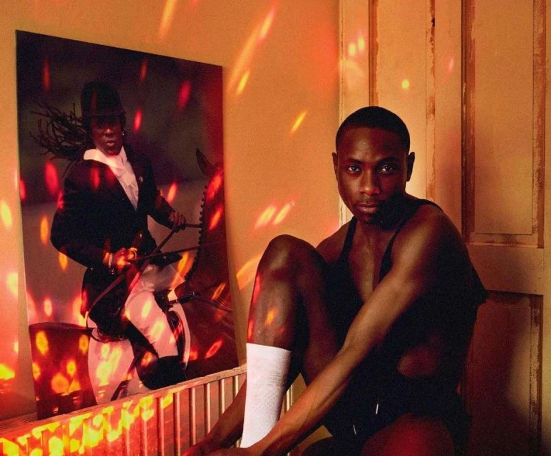 Portrait of a Black man in a tank top sitting in front of a framed image of a person riding a horse, illuminated by red and orange light.