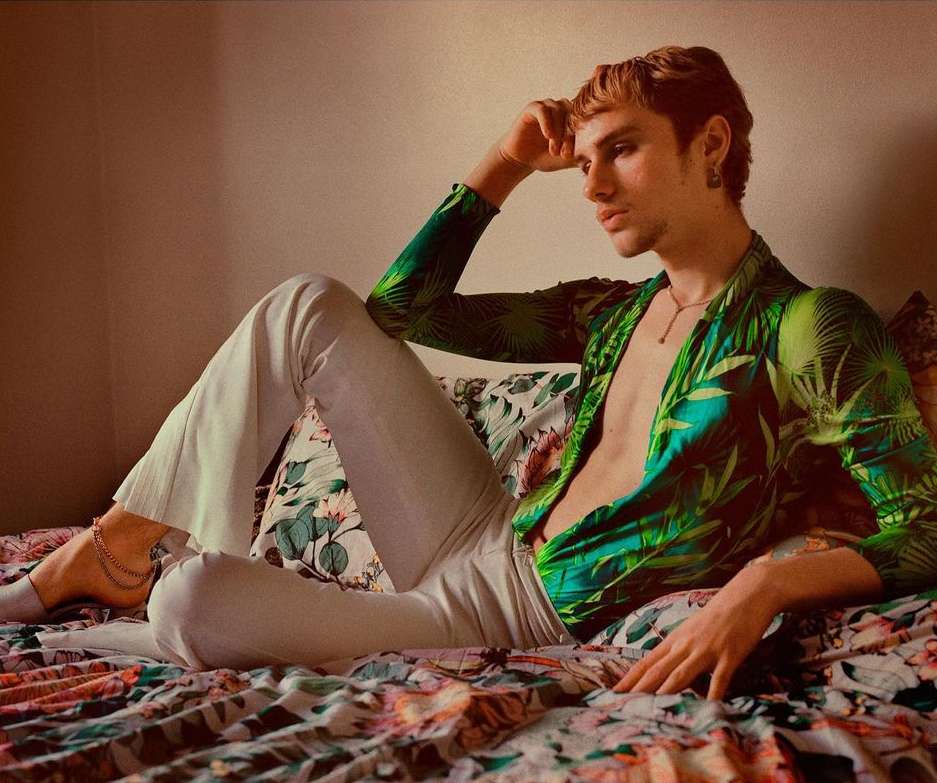 A blond male model poses on a bed in a green tropical-print shirt and white pants, with a soft, warm-toned aesthetic, evoking 70s fashion.