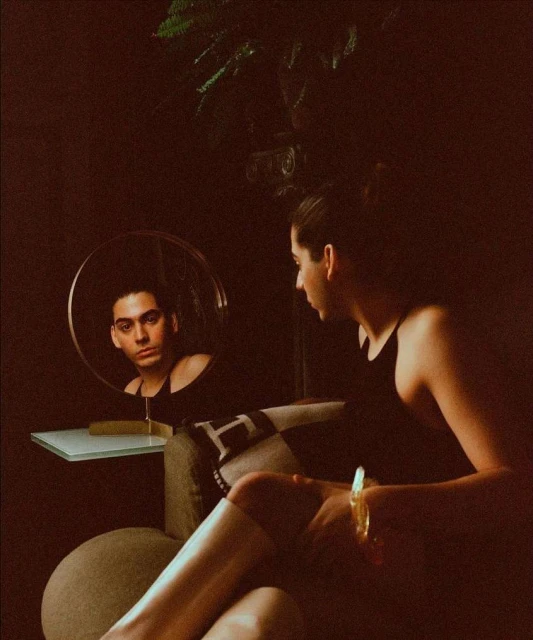 Woman seated, leg visible, reflected in round mirror, revealing a man's face; dark indoor setting with minimal visible lighting.