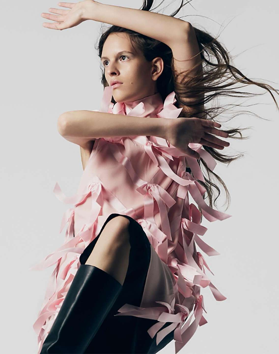 Fashion model poses in a pink ribbon dress and black boots against a neutral backdrop in a studio setting with wind-blown hair.