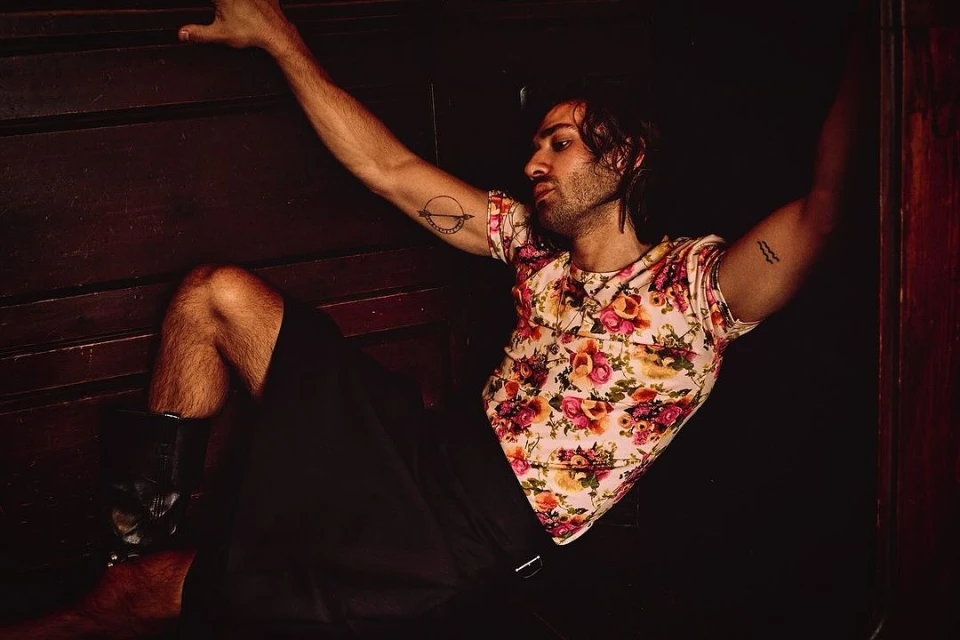 Man in floral shirt poses indoors with arm raised, legs bent, and boots. Dark, moody lighting emphasizes tattoos and casual attire.