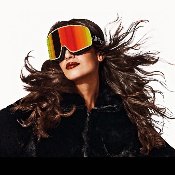 Brunette woman poses in a black fur jacket and ski goggles with red, orange, and gold lenses against a white studio backdrop.