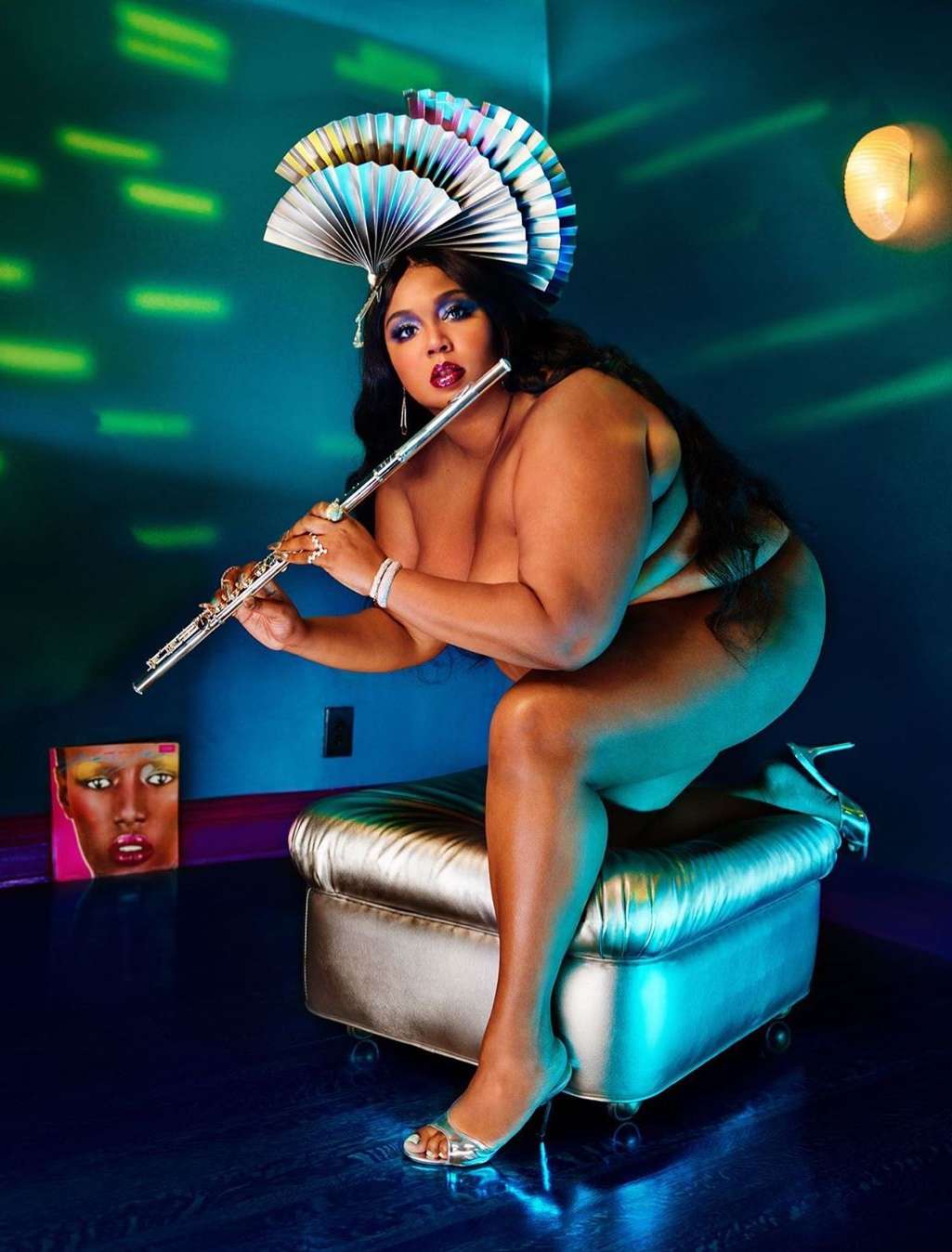 Studio shot of bare-shouldered woman playing flute on metallic stool. She wears a fan-shaped headpiece and high heels, and faces the camera.