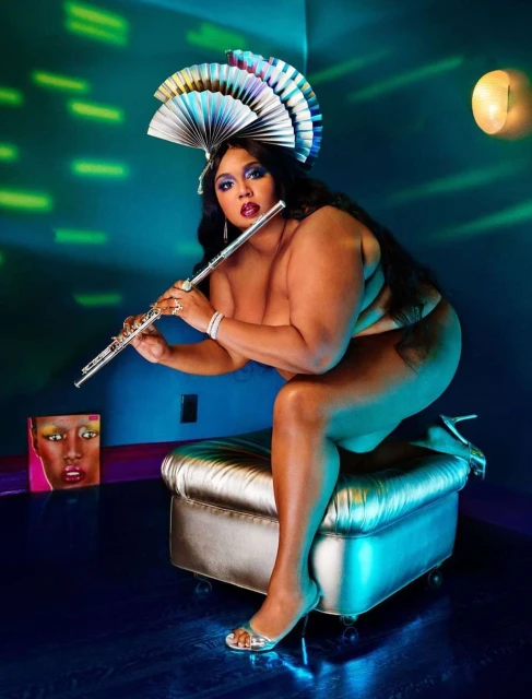 Studio shot of bare-shouldered woman playing flute on metallic stool. She wears a fan-shaped headpiece and high heels, and faces the camera.