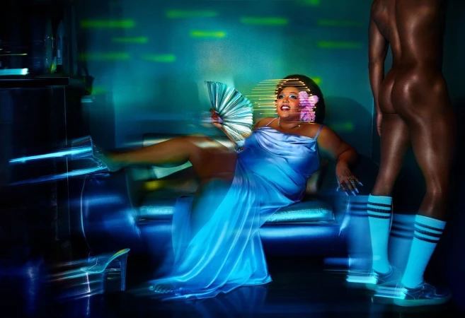 Woman in satin blue dress reclining on sofa holding fan with standing male figure