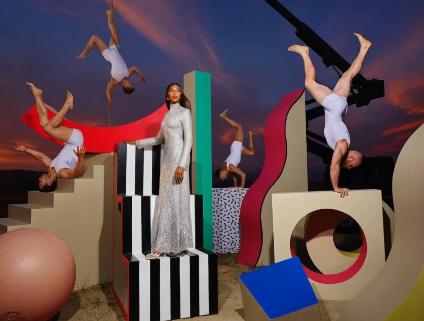 Fashion editorial shot with a model in a sequined gown, gymnasts in white leotards, and colorful geometric props against a twilight sky.
