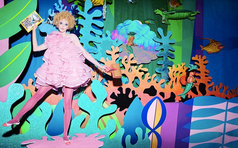 Young woman in pink dress and matching tights stands in a playful set of cardboard mermaids, coral, and sea plants holding book and container.