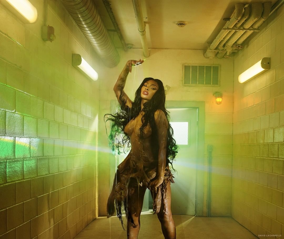 A dark-haired woman poses with water dripping on her in a tile-lined interior. Moody, low-key lighting, and earth-toned color palette.