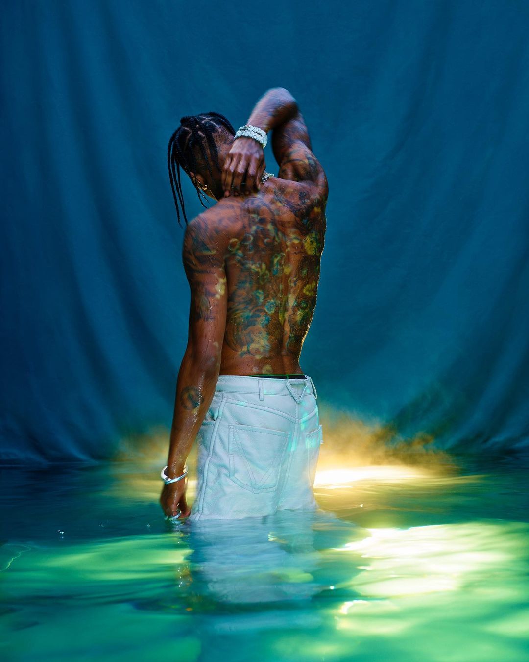 Shirtless African-American male partially submerged in water with colorful tattoos on his back against a blue backdrop.