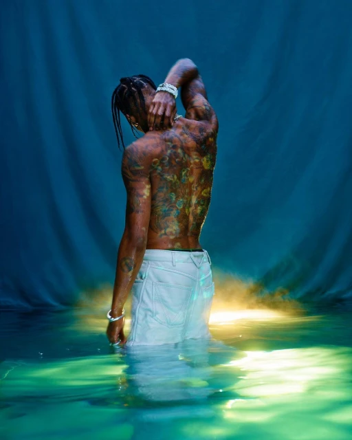 Shirtless African-American male partially submerged in water with colorful tattoos on his back against a blue backdrop.