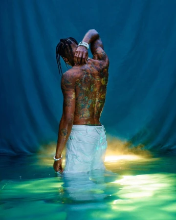 Shirtless African-American male partially submerged in water with colorful tattoos on his back against a blue backdrop.