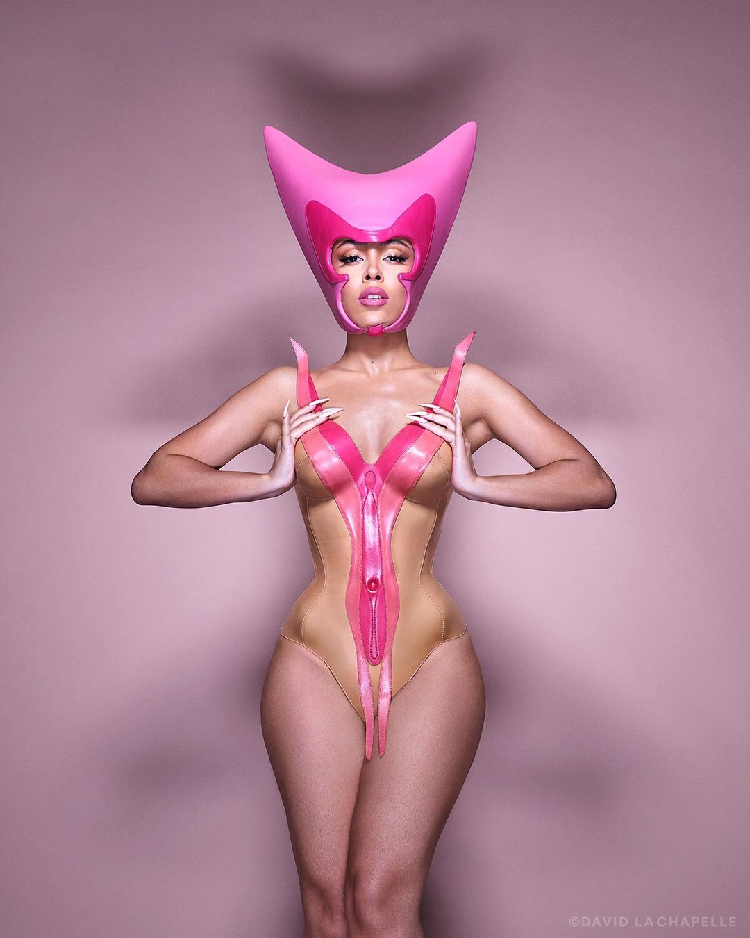Adult female posing with creative pink headwear and bodysuit in a pink photo studio. Studio lighting and fashion forward design.