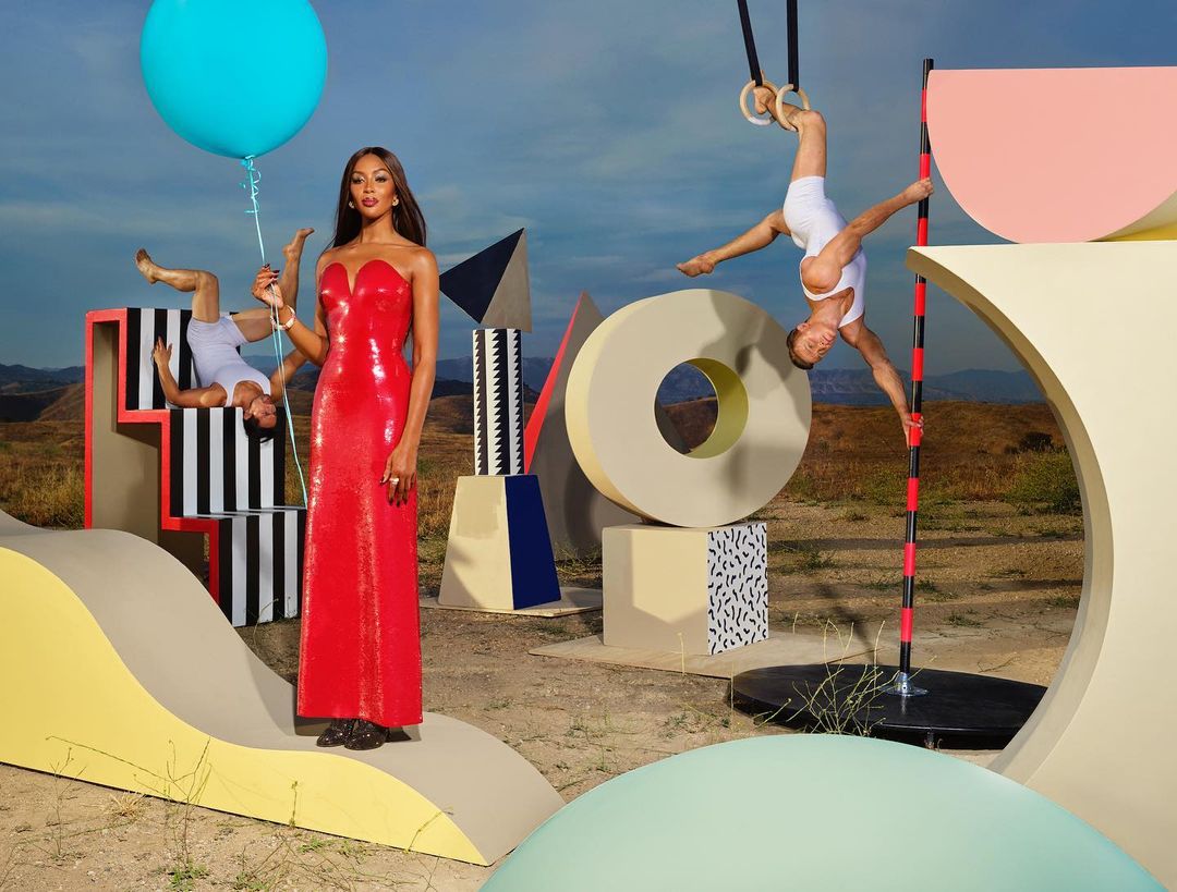 Fashion shot of Naomi Campbell in red dress holding a blue balloon, surrounded by geometric forms and a man on gymnastic rings.