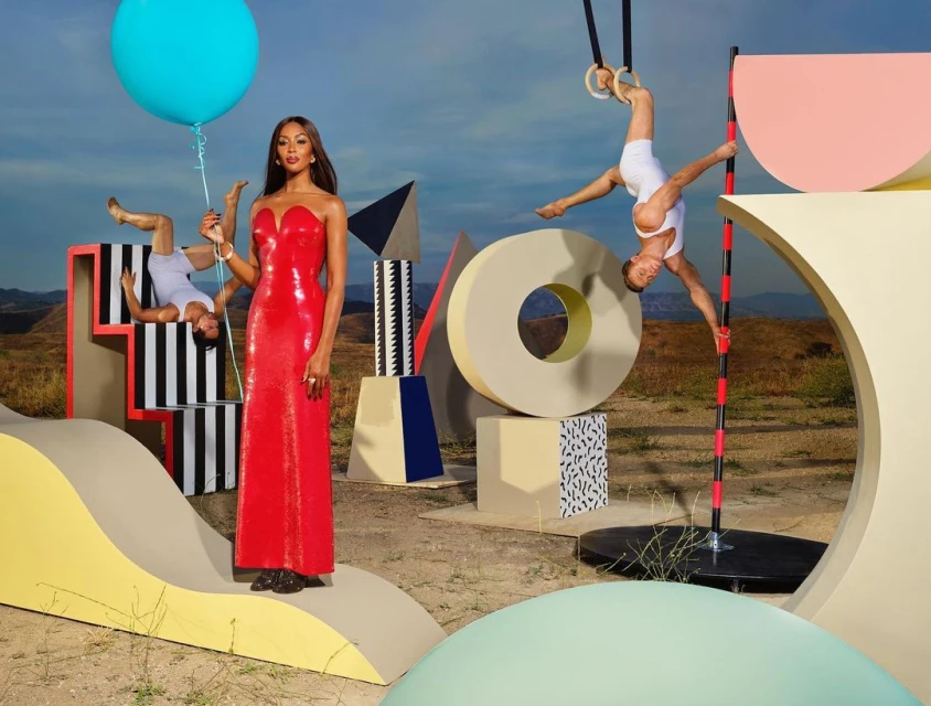 Fashion shot of Naomi Campbell in red dress holding a blue balloon, surrounded by geometric forms and a man on gymnastic rings.