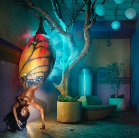 Artistic portrait of a woman bending backwards beneath a butterfly wing-shaped lamp, with indoor plant and minimalist interior design.