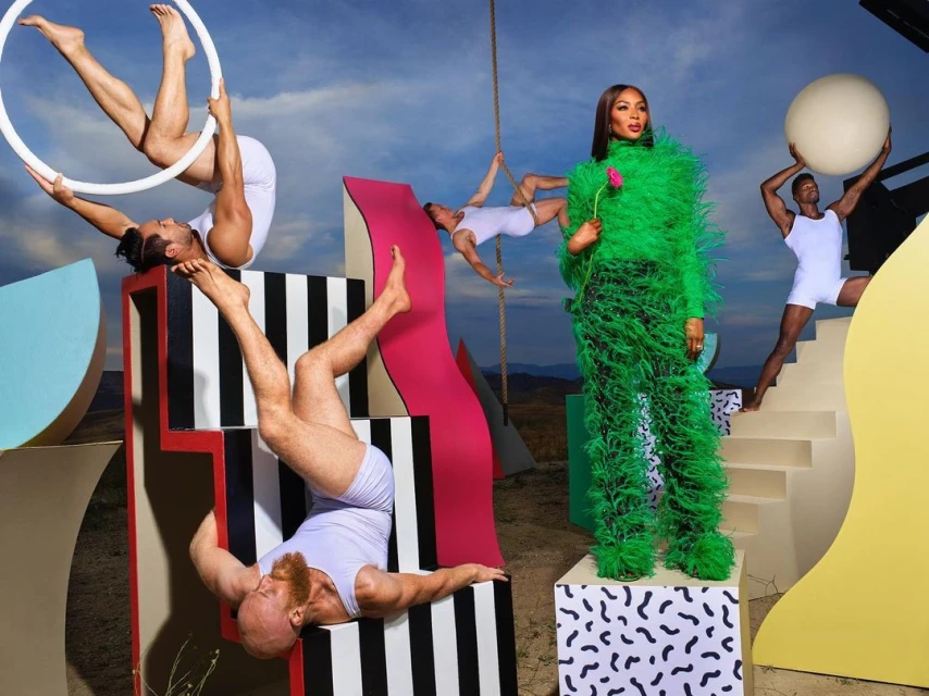 Fashion editorial shot with a woman in a green feathered suit posing with a flower, surrounded by male dancers in a desert setting.