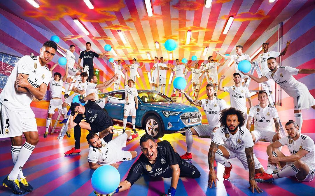 Promotional shot of Real Madrid soccer team and blue Audi e-tron car in colorful studio setting with blue balls and white and orange background.