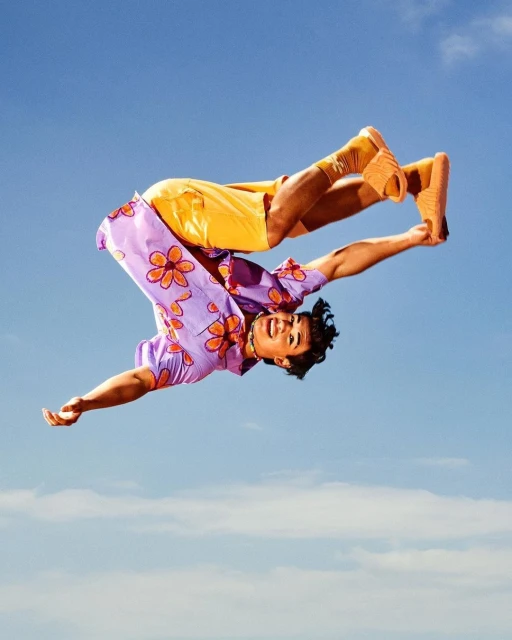 Young man in mid-air performing a backflip against a blue sky wearing a purple floral shirt, yellow shorts, and orange slides.