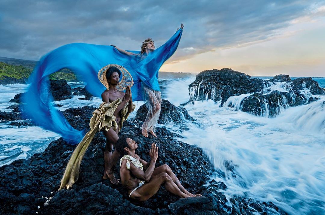 Three models in elegant drapery pose on lava rocks by ocean waves under a cloudy sky, evoking high-fashion editorial and nature themes.