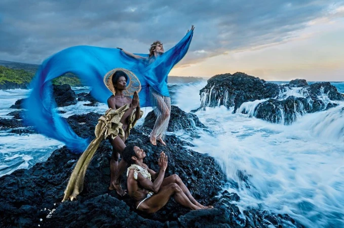 Three models in elegant drapery pose on lava rocks by ocean waves under a cloudy sky, evoking high-fashion editorial and nature themes.
