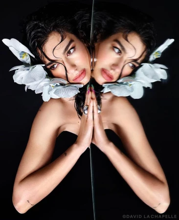 Close-up studio portrait of a woman with dark hair, adorned with white orchids, posing with hands in front of a mirror reflection.
