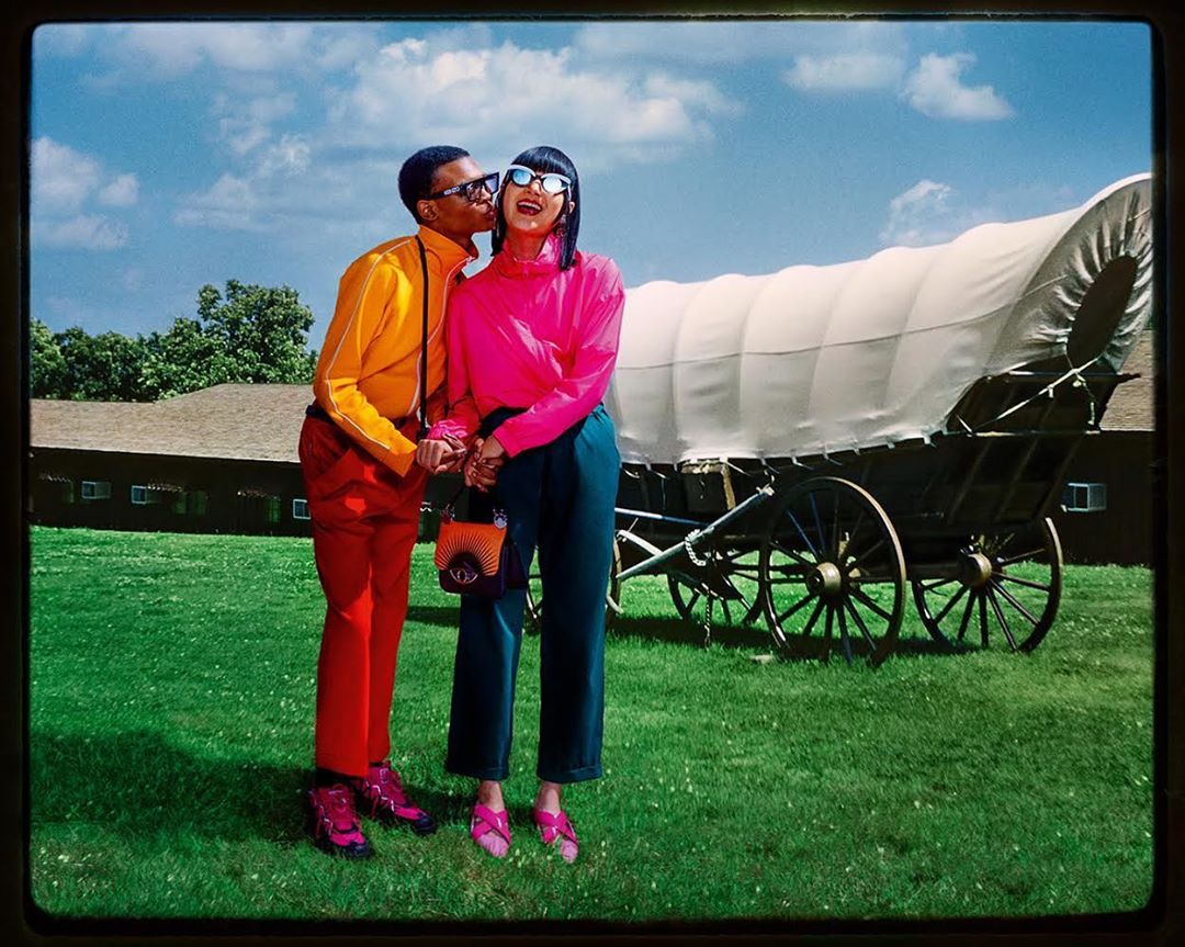 A man kisses a woman on the cheek in a field in front of a covered wagon, both wearing bright clothing and sunglasses.