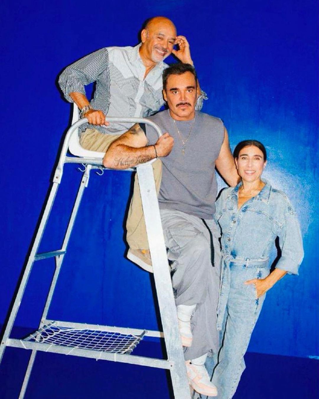 Three adults pose in front of a blue backdrop: two men, one on a ladder, and one woman wearing a denim jumpsuit.