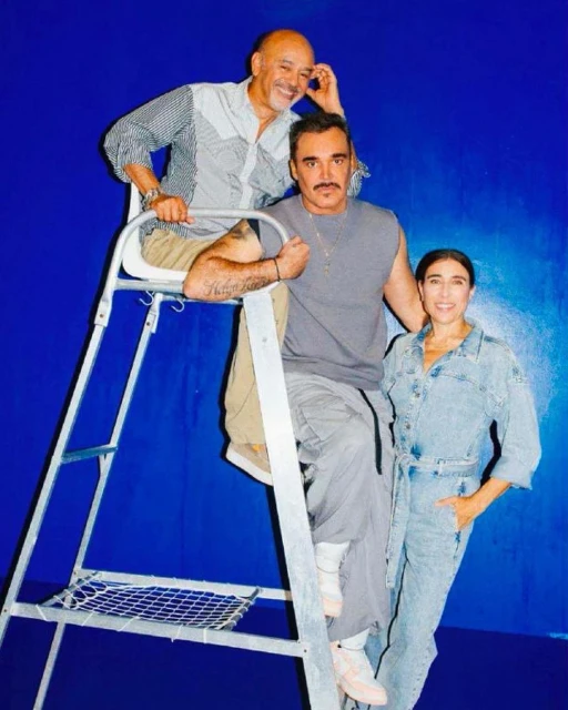 Three adults pose in front of a blue backdrop: two men, one on a ladder, and one woman wearing a denim jumpsuit.