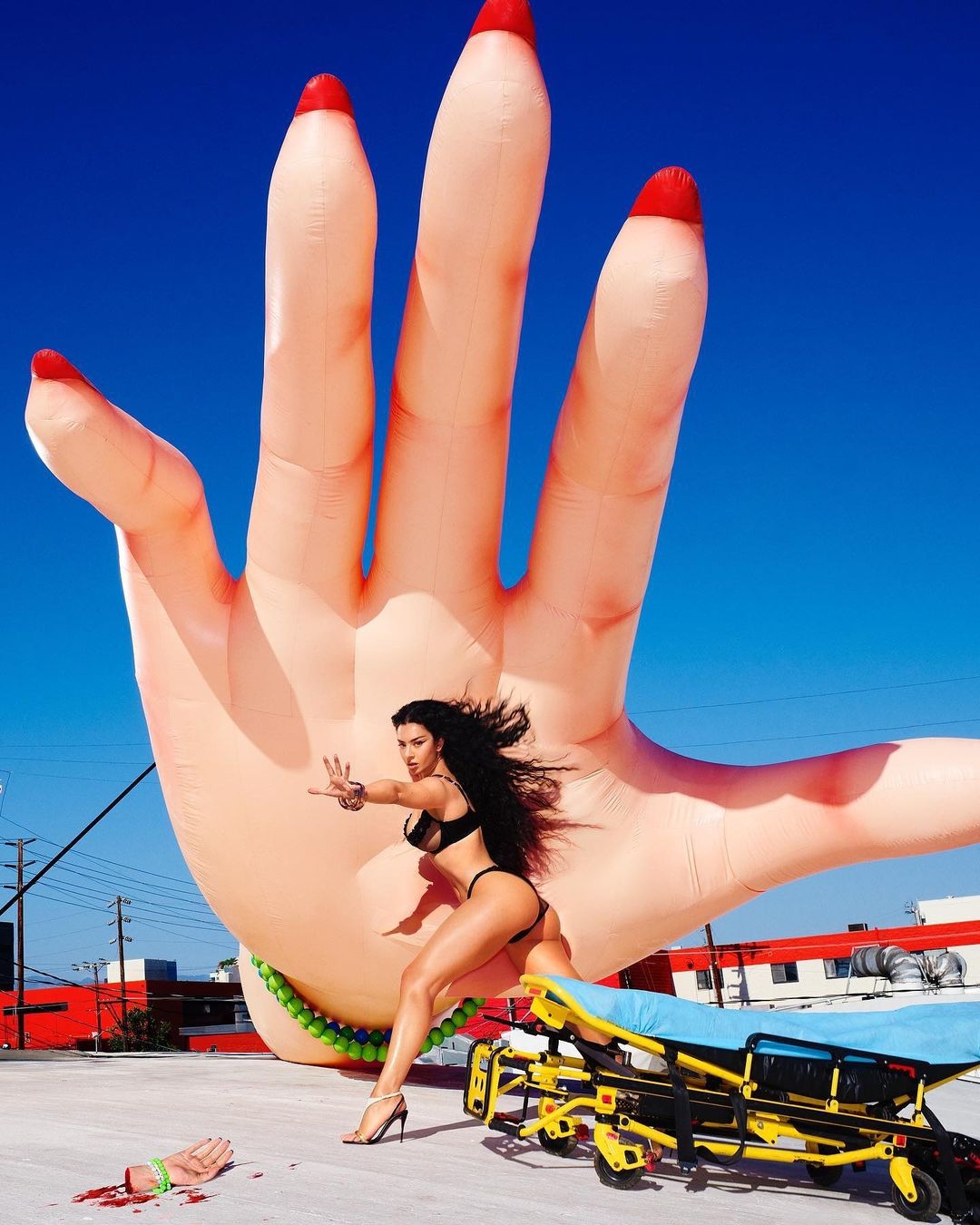 A woman in a black bikini poses in front of an inflatable hand on a rooftop next to a yellow gurney against a blue sky backdrop.
