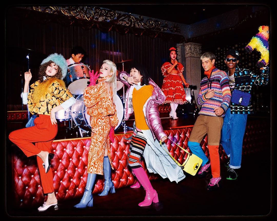 Group of diverse adults in colorful retro-inspired clothing posing in a vibrant, indoor performance space with stage lights and musical instruments.