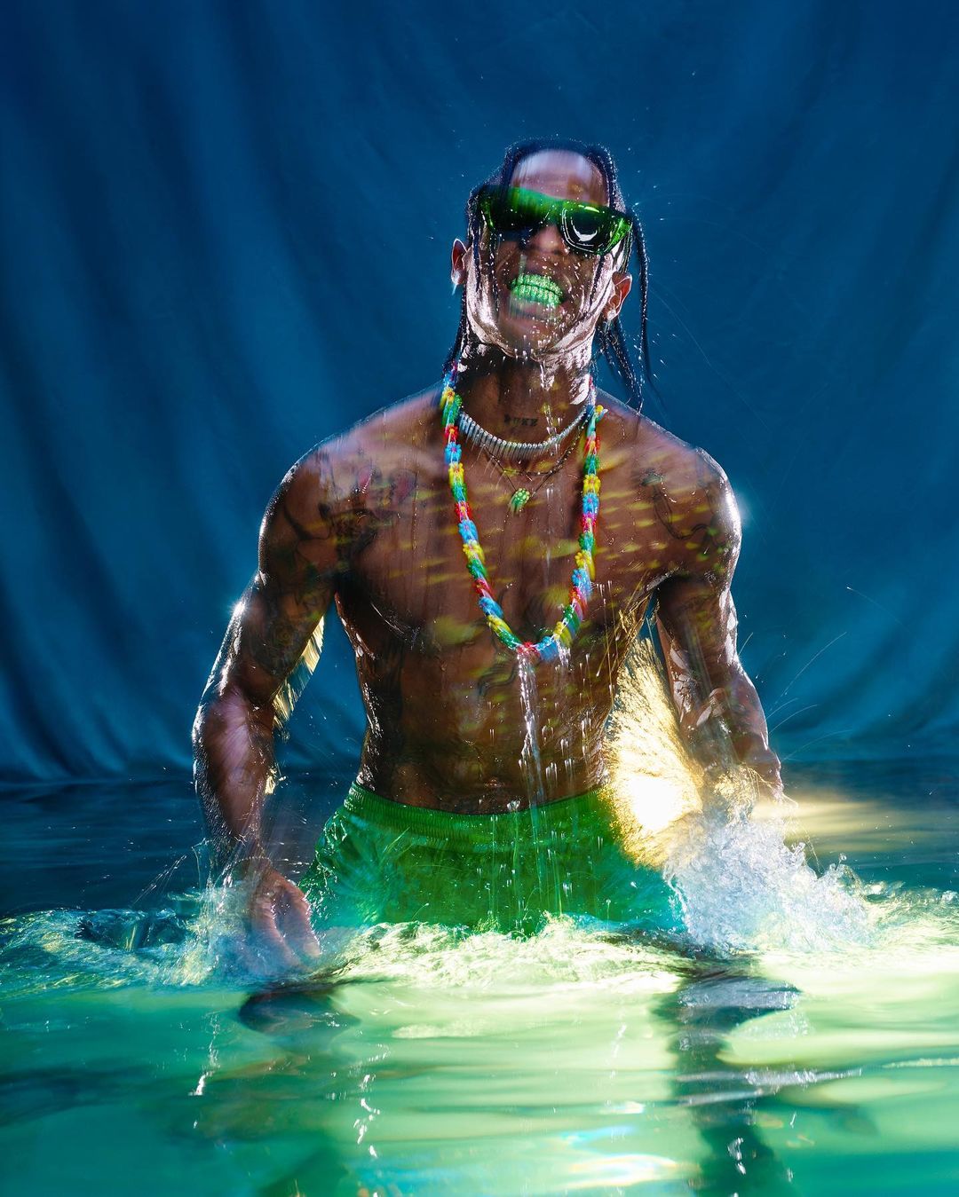 Adult male standing in water with green accents, wearing sunglasses and a colorful necklace, against a teal background. 