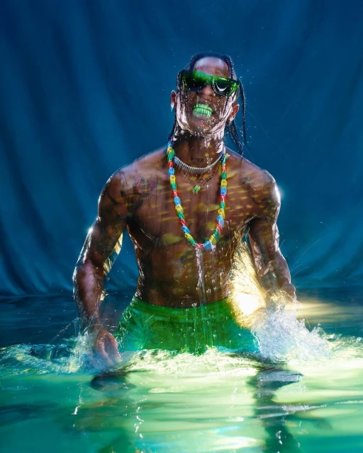 Adult male standing in water with green accents, wearing sunglasses and a colorful necklace, against a teal background. 
