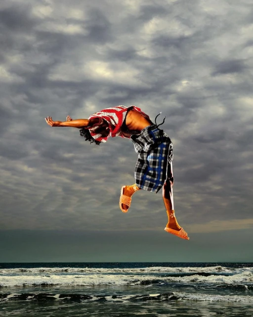 Adult male mid-air backflip on ocean shore with red patterned shirt, plaid shorts, and sandals, against a cloudy sky backdrop.