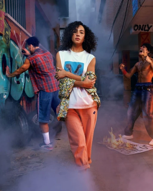 A young woman stands confidently in an urban alleyway with graffiti, two men, smoke effects, and a burning protest sign visible.
