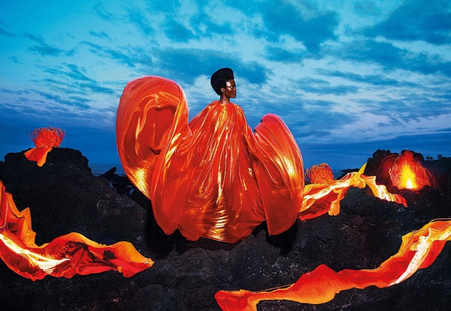 An African woman poses on a rocky landscape in a flowing red dress with glowing lava, set against a blue sky with scattered clouds.