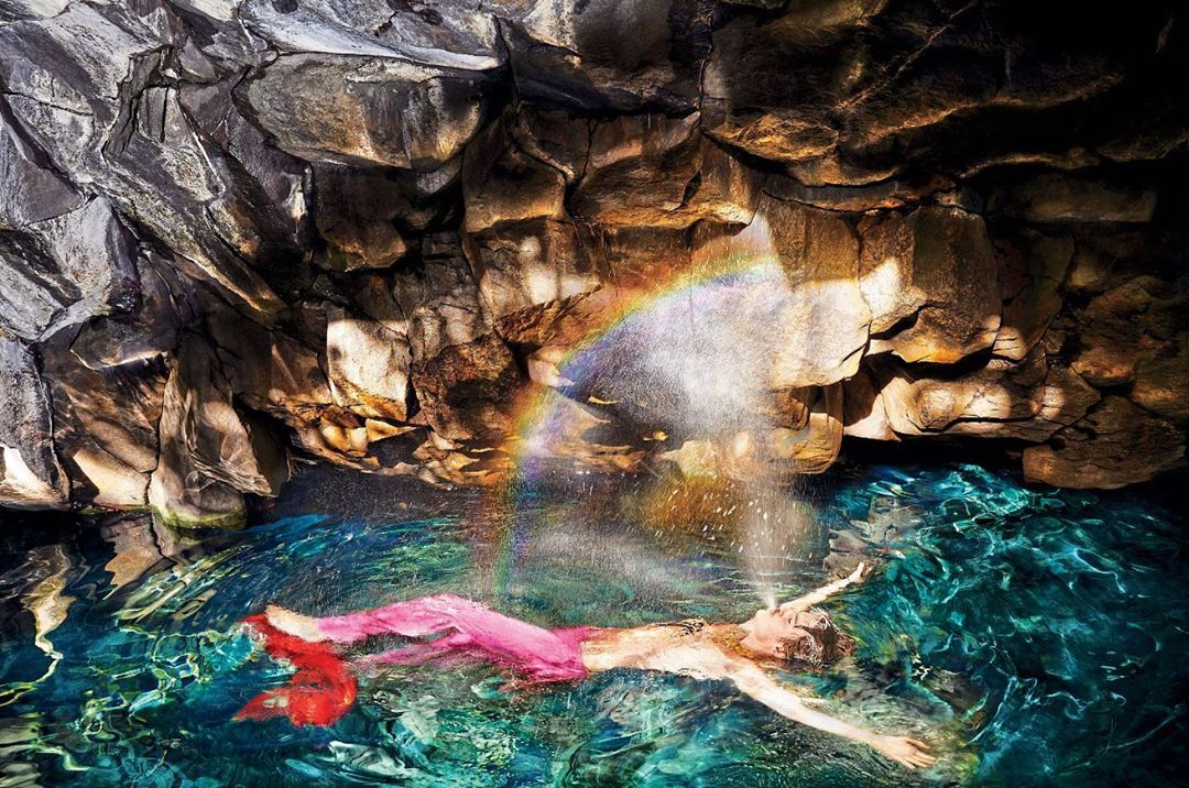 Woman floats in turquoise water of a rocky cave with rainbow light casting colours across the scene, creating an ethereal effect.