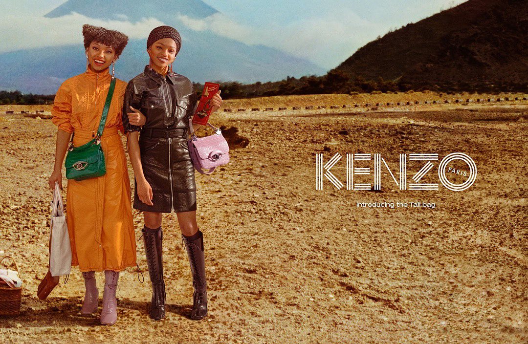 Two women pose with Kenzo Paris bags in an outdoor fashion shot. A mountain rises in the background. The Kenzo logo is in view.