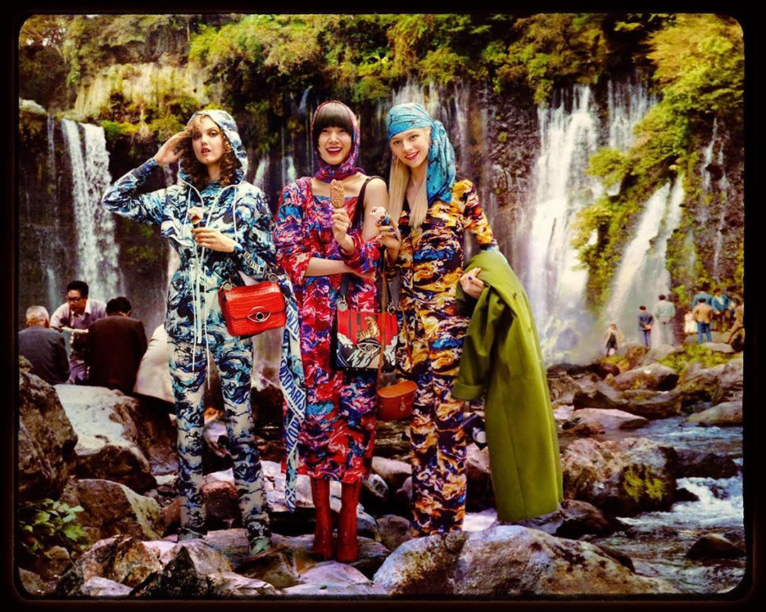 Three models in patterned jumpsuits pose on rocks near a waterfall. They are holding snacks and purses and are wearing headscarves.