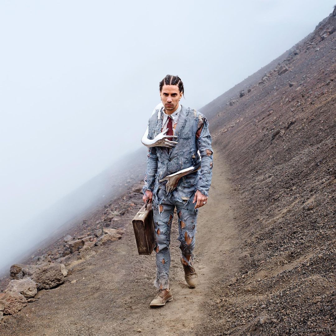 Man in tattered suit walks path on volcanic slope with suitcase. Fog obscures the background. Conceptually rich in symbolism.