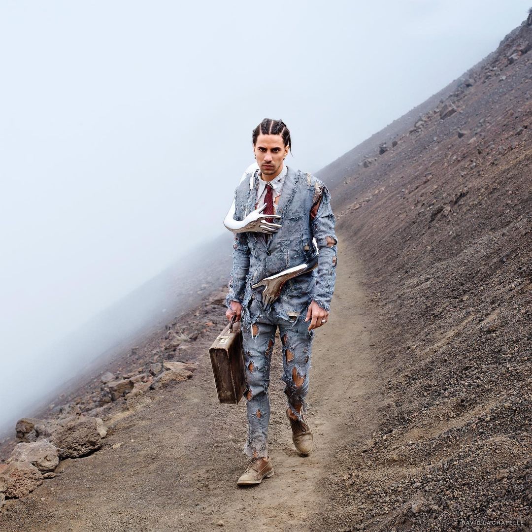 A man in a tattered suit and tie carrying a briefcase walks on a rocky path. A landscape with fog and a mountain in the background.
